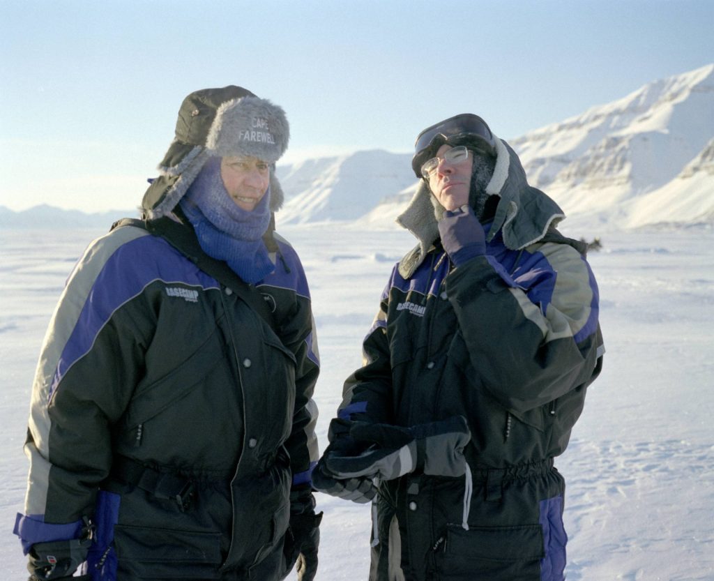 L'artista David Buckland e l'autore Ian McEwan conversano durante la spedizione artica del 2005 compiuta per il progetto Cape Farewell. Indossano vestiti pesanti invernali, adatti per le basse temperature artiche. Stanno in piedi su un terreno innevato e desolato e in sottofondo si vede una montagna di neve. C'è la luce naturale del sole.